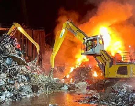 Heavy machinery tackling a large industrial fire involving mixed scrap materials and potential lithium battery hazards.